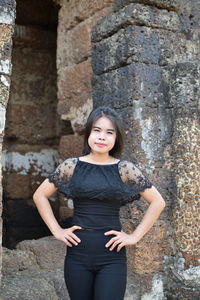 Portrait of young woman with hands on hip standing against old ruin temple