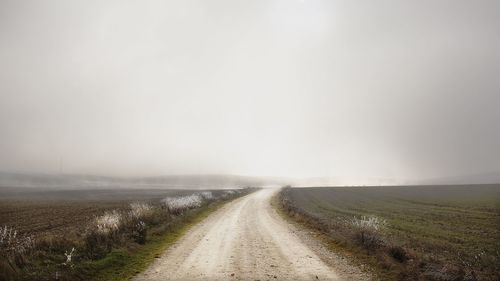 Dirt road amidst field against sky during foggy weather