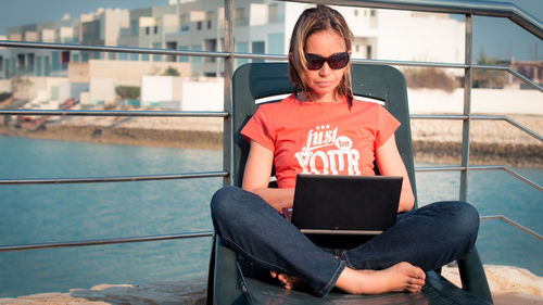 Woman working on laptop while siting on deck chair against railing
