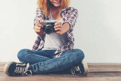 Young woman photographing from camera while sitting against wall on wooden floor