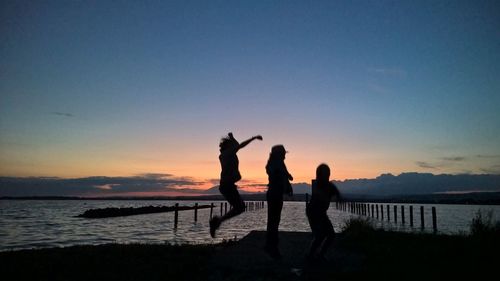 Silhouette siblings enjoying on lakeshore against sky during sunset