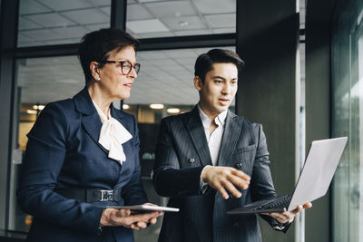 Young businessman discussing over laptop with senior coworker while standing in office