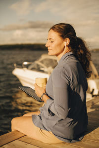 Smiling mature woman having coffee while using digital tablet at patio against lake during vacations