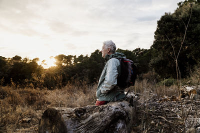 Senior male hiker with hands in pockets sitting on wooden log while looking at view during weekend