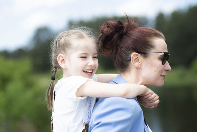 Happy little girl sitting on her mother's shoulders or back in sunny summer day