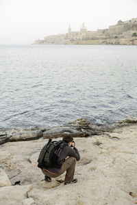 Photographer takes picture of something while kneeling on rock by sea. man travels around malta isla