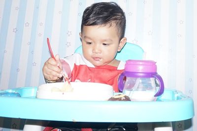 Close-up of cute baby boy eating food while sitting on high chair against wall 