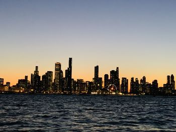 Sea by modern buildings against clear sky during sunset