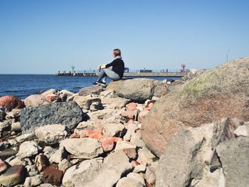 Man sitting on rock by sea against clear sky