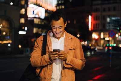 Smiling young man texting through phone while standing in illuminated city at night