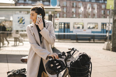 Businesswoman talking on smart phone standing with bicycle while looking back over shoulder in city