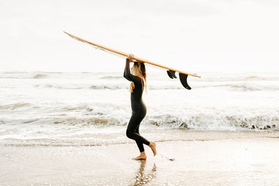 Side view of female surfer dressed in wetsuit walking looking away while holding surfboard on head on the beach during sunrise in the background