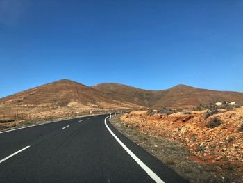 Road leading towards mountains against clear blue sky