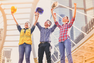 Low angle view of architectures with arms raised holding hardhats while standing against building