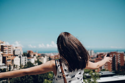 Rear view of woman with arms outstretched looking at cityscape against sky