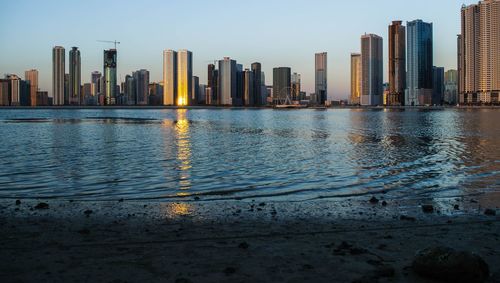 Modern cityscape by sea against sky during sunset