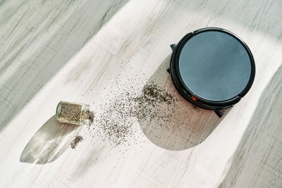 From above of round robotic vacuum cleaner sliding on light laminate floor and removing dirt spilled out of glass pot