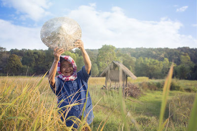 Portrait of farmer holding asian style conical hat on field against sky