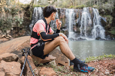Side view of male hiker with hydration pack drinking from hose while having break during trekking in forest and admiring view of lake near waterfall
