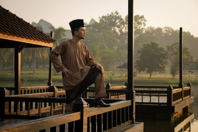 Man wearing traditional clothing while standing on built structure against lake during sunset