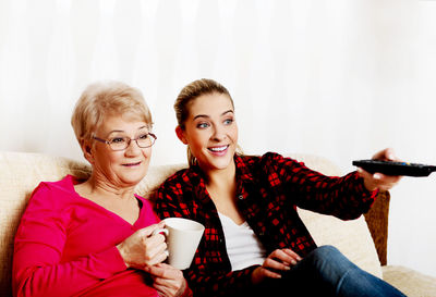 Smiling woman holding remote control while sitting with senior woman on sofa at home