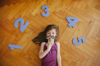 Directly above shot of girl holding artificial mustache and numbers while lying on floor
