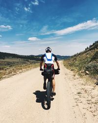 Rear view of man cycling on dirt road against sky