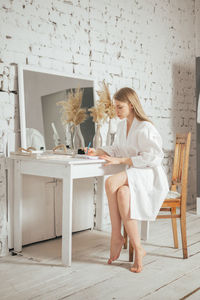 Young blond woman at home wearing white bathrobe writing down notes at paper notebook white palette