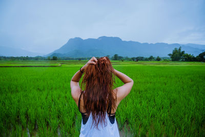 Rear view of young woman with hand in hair standing on field against sky