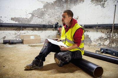 Male plumber reading document while sitting on floor by pipes at basement