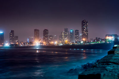 Illuminated buildings by sea against sky at night