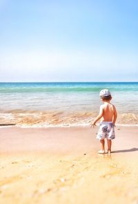Rear view of shirtless boy standing on shore at beach against sky during sunny day