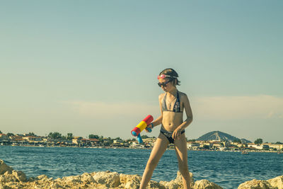 Girl wearing bikini at sea by rocks against sky