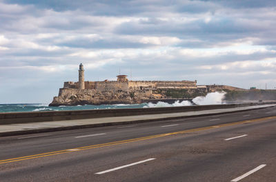 Road by buildings against cloudy sky