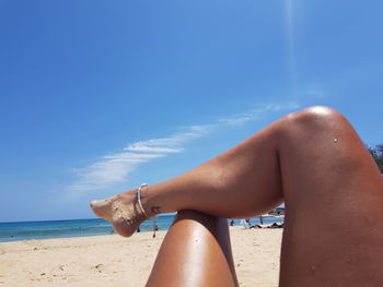 Low section of woman relaxing on beach against blue sky