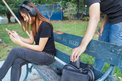 Midsection of man removing wallet from bag while woman using phone on bench