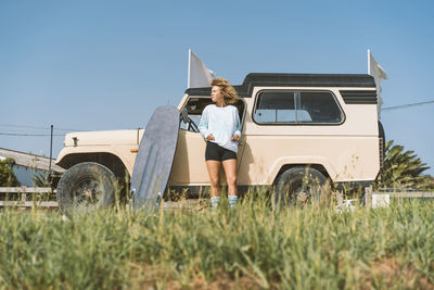 Young afro woman looking away while standing with surfboard against old off-road vehicle on sunny day