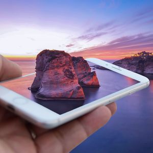 Digital composite image of person holding mobile phone with mountain against sky during sunset