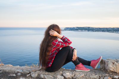 Young woman sitting by sea against sky