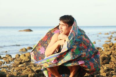 Man wrapped in blanket crouching on rocky shore