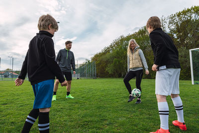 Happy sporty couple and teenage boys playing football together while kicking ball in green field in summer