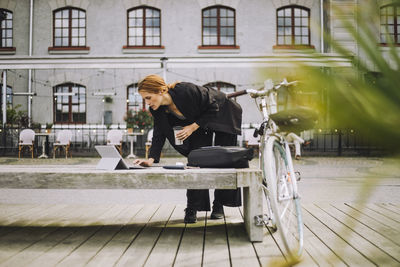 Female freelancer holding insulated drink container using laptop on bench at promenade