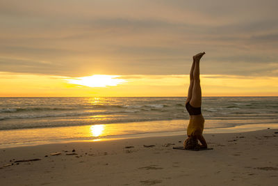 Woman doing headstand at beach against sky during sunset