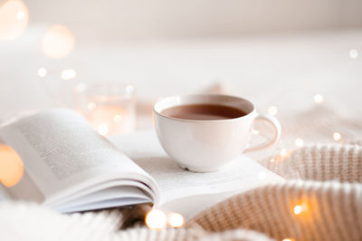 Open book with cup of coffee in bed with knit cloth sweater over glow christmas lights at background 