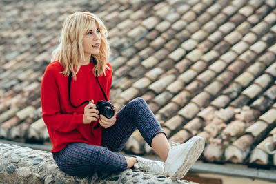 Beautiful woman with camera sitting on concrete wall by roof 