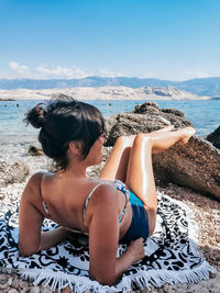 Rear view of beautiful young woman in bikini sunbathing on rocky beach.