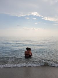 Rear view of mother sitting with son in sea against cloudy sky