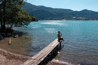 Mid adult man with beer bottle sitting on pier over lake against clear blue sky