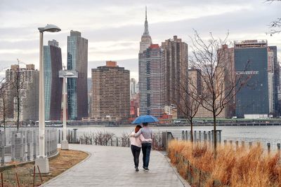 Rear view of couple walking with umbrella on road by river and buildings