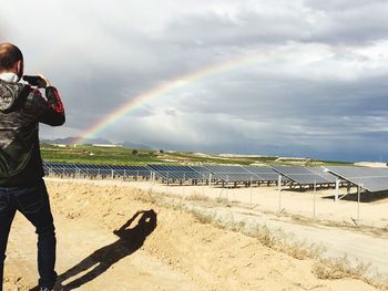 Man photographing rainbow while standing against cloudy sky